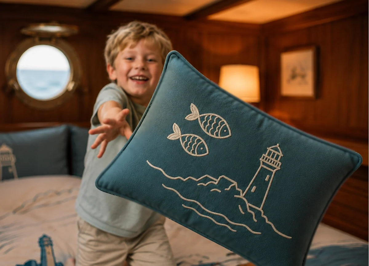 A smiling young boy in a berth on a sailboat with wood-paneled walls holds up a teal blue cushion featuring a cream-colored embroidered fish and the FastNet Rock lighthouse design.