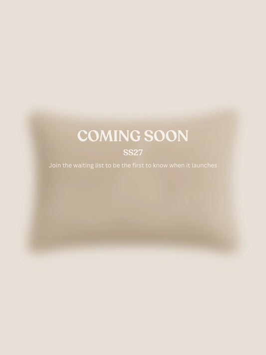 A soft beige rectangular cushion shown on a light warm beige background with centered text reading 'COMING SOON' and smaller text 'SS27 Join the waiting list to be the first to know when it launches.'.