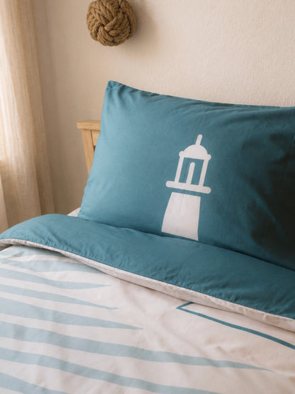 A teal blue pillowcase with a white lighthouse illustration, shown on a bed with a matching blue and white duvet cover in a softly lit bedroom.