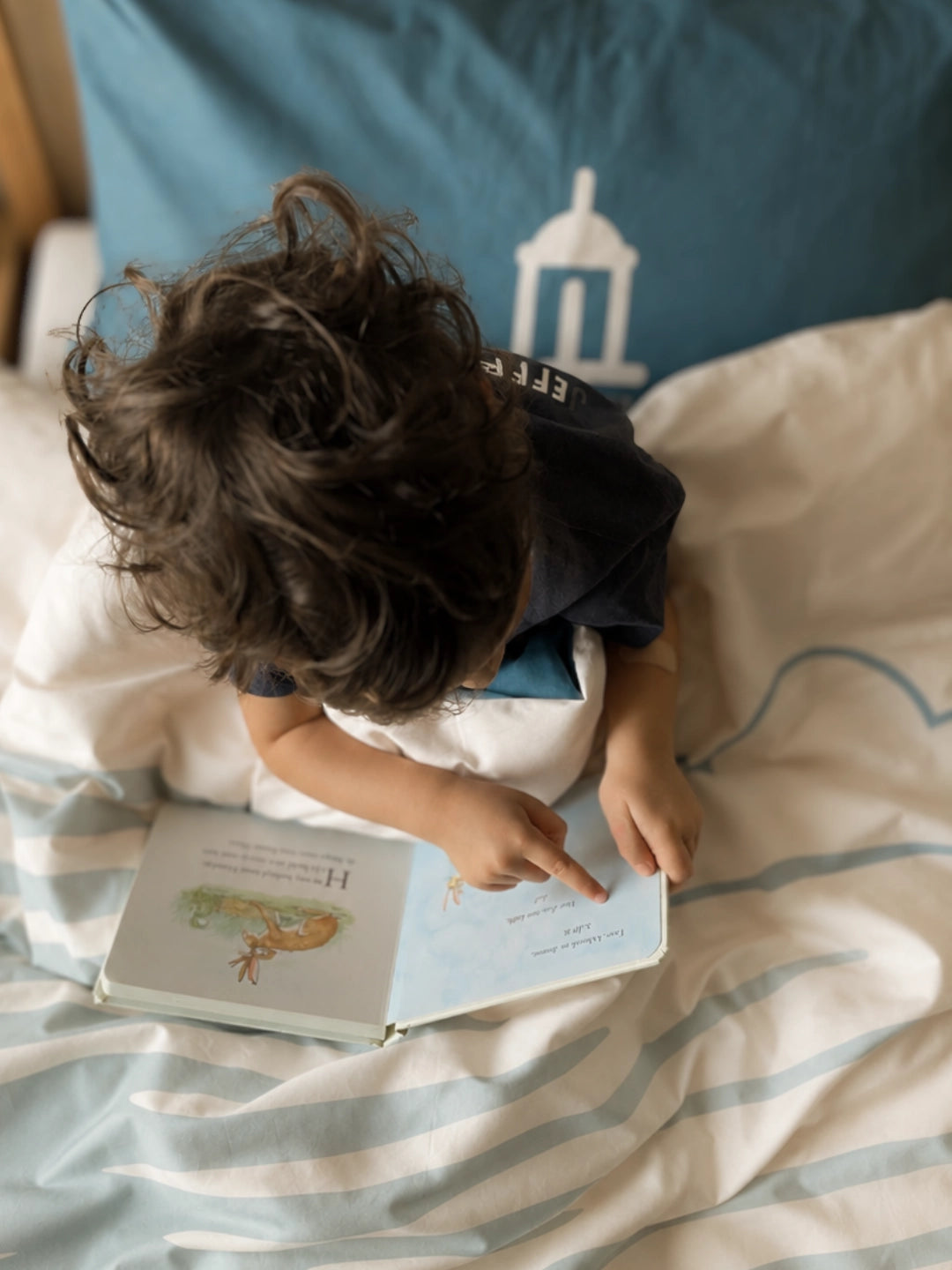 A toddler with curly dark hair sits on a bed reading a children's book, pointing at the page. The bed has white and blue bedding with a blue pillow featuring the FastNet Rock lighthouse.