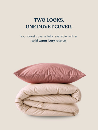 A duvet cover set set shown on a warm beige background with marketing text. The top pillow is dusty rose, and the folded duvet underneath is a warm ivory, illustrating the reversible nature of the product.