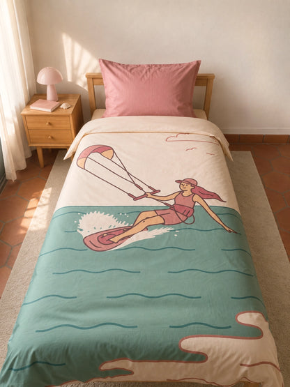 A single bed in a softly lit bedroom, styled with an illustrated organic cotton duvet cover for kids featuring an illustrated girl Kitesurfing on a teal ocean scene, with a soft pink pillow and a small wooden bedside table with a pink lamp.