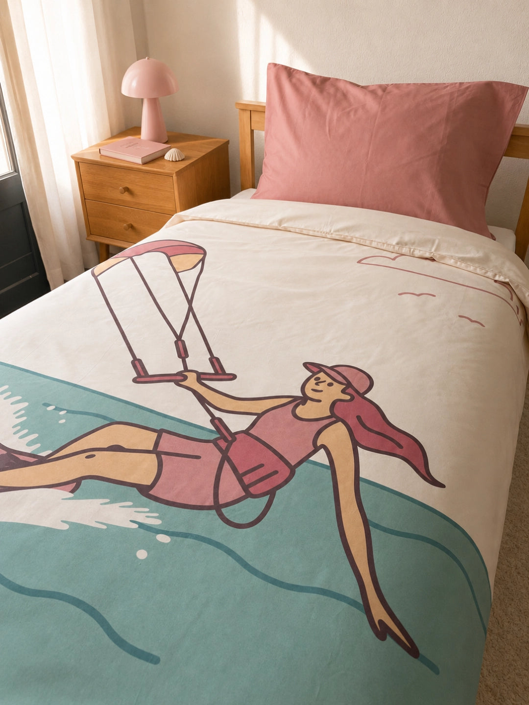 A single bed in a bedroom setting with an organic cotton duvet cover for kids featuring a retro-style illustration of a girl Kitesurfing. The duvet is off-white with a large teal wave at the bottom and a soft pink pillowcase.