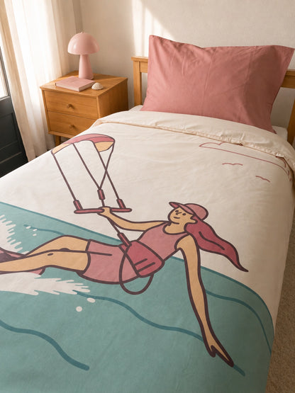 A single bed in a bedroom setting with an organic cotton duvet cover for kids featuring a retro-style illustration of a girl Kitesurfing. The duvet is off-white with a large teal wave at the bottom and a soft pink pillowcase.