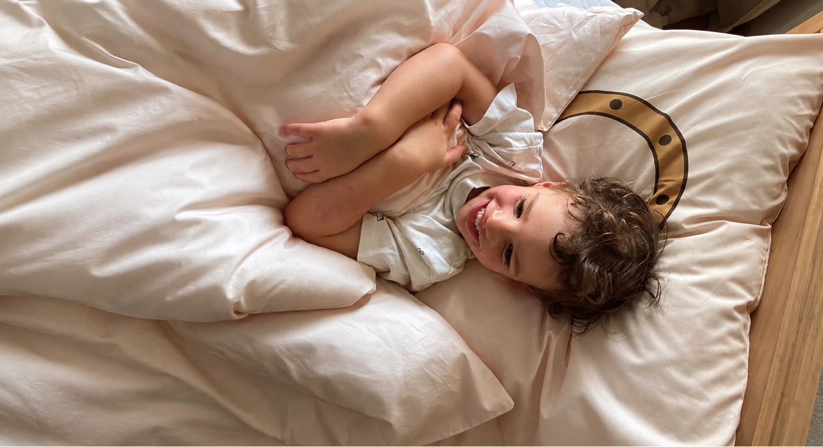 A smiling toddler lies in bed under a soft cosy organic cotton duvet cover with a matching pillow.