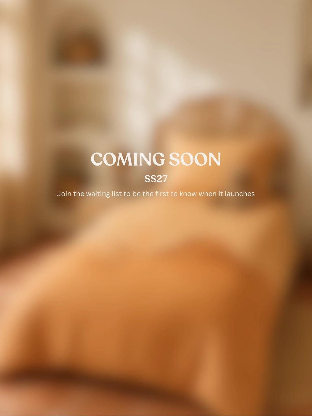 A blurred warm-toned interior scene showing a bed with orange bedding, with overlaid text reading 'COMING SOON SS27 Join the waiting list to be the first to know when it launches'.