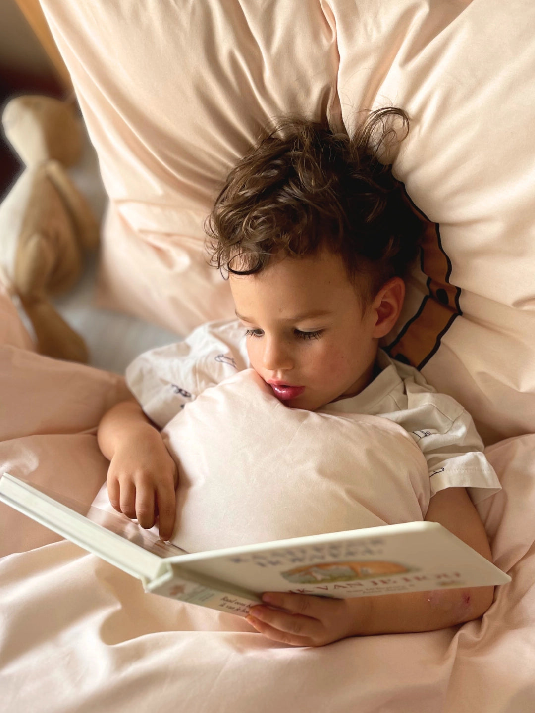 A toddler with curly brown hair lies on soft organic cotton bedding, holding and reading a book. 