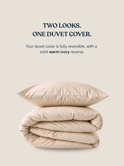 A folded beige organic cotton duvet cover on a warm cream background, with text above reading 'TWO LOOKS. ONE DUVET COVER.