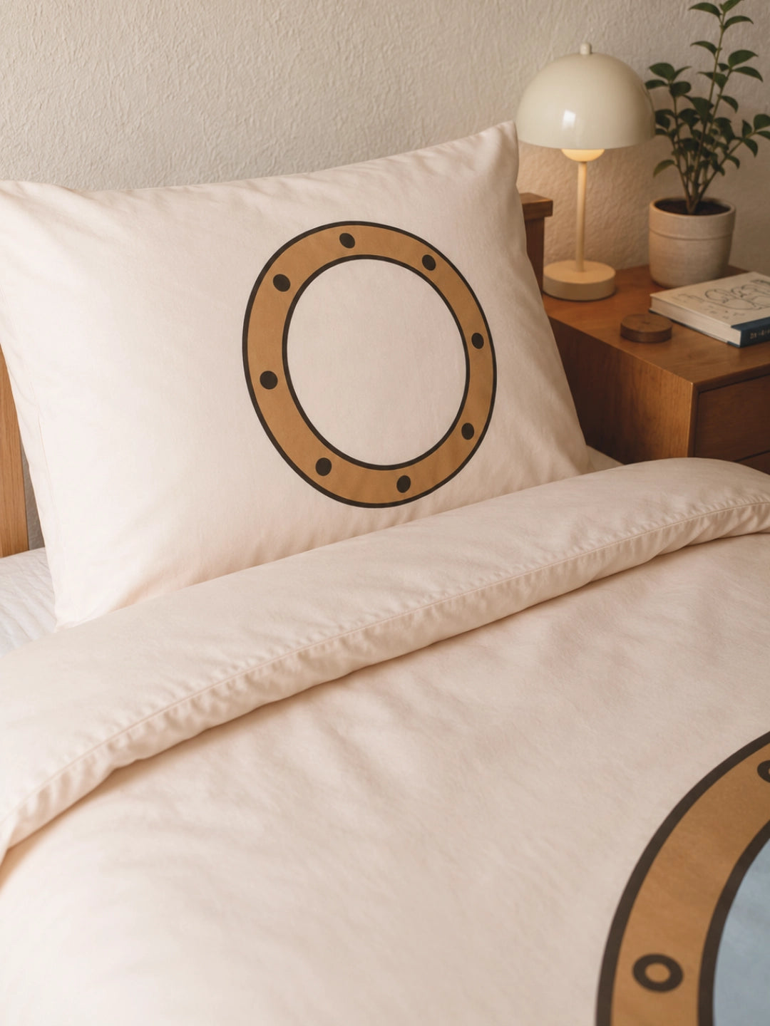 A close-up of an organic cotton duvet cover set with a porthole graphic printed on the pillow and matching duvet cover. A wooden nightstand with a white lamp and small plant is visible beside the bed.