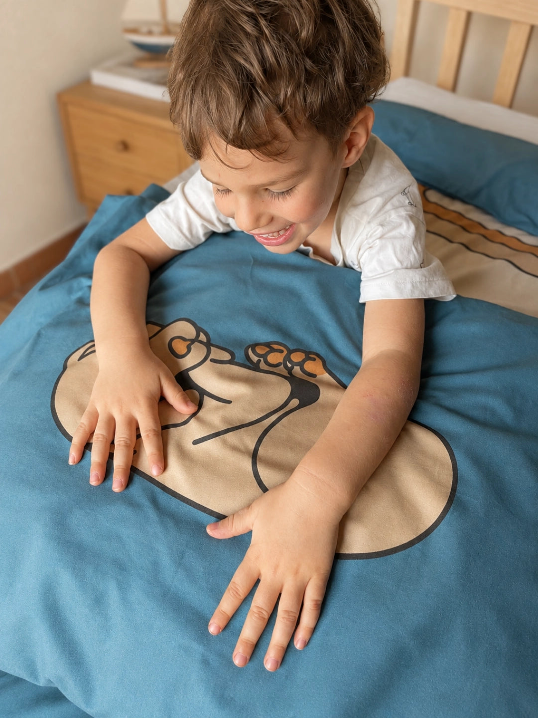 A toddler with brown hair lies on a bed, smiling  with his hands on a blue pillowcase illustrated with a beige dog. 