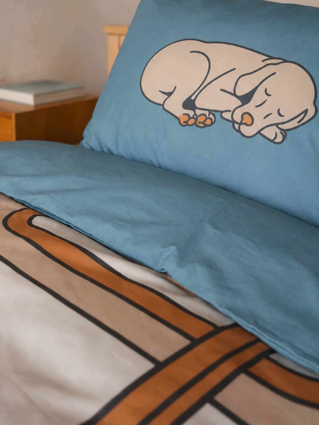 A close-up of a single bed with a light blue pillowcase featuring a sleeping dog illustration, paired with a matching duvet cover.