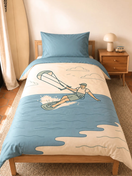 A single bed dressed in a light blue and cream duvet cover with a vintage-style illustration of a Kitesurfer, shown in a warm, neutral-toned bedroom setting.